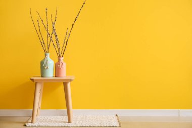 Vases with willow branches on table near yellow wall