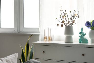 Vases with tree branches, Easter eggs, flowers and rabbits on chest of drawers in room