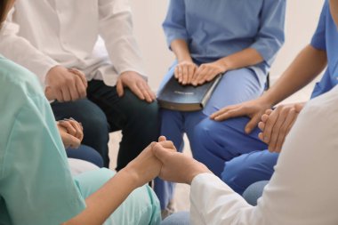 Group of doctors praying with Holy Bible in clinic, closeup