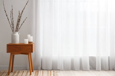 Vase with willow branches and candles on table near light curtain