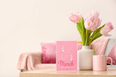 Vase with tulips, cup and greeting card for Women's Day on table in living room