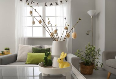 Vase with tree branches, Easter eggs and rabbits on table in light living room