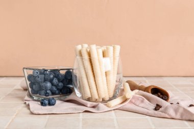 Glass of delicious wafer rolls and bowl with blueberries on tile table