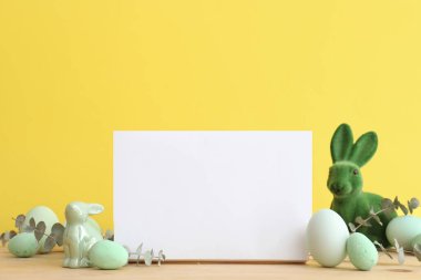 Blank frame, Easter bunny, painted eggs and eucalyptus leaves on wooden table near yellow wall