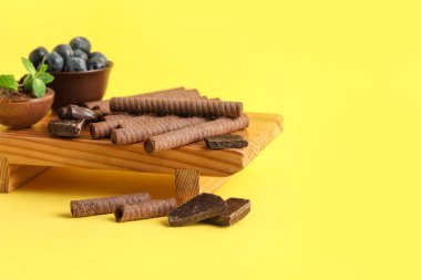 Board with delicious wafer rolls, chocolate pieces and blueberries on yellow background