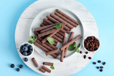 Board with delicious chocolate wafer rolls, blueberries and coffee beans on blue background