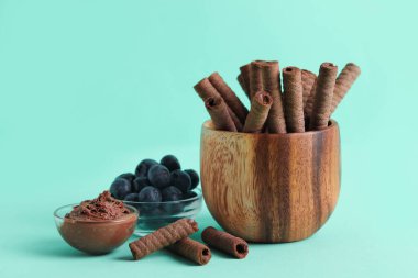 Bowls with delicious chocolate wafer rolls and blueberries on turquoise background