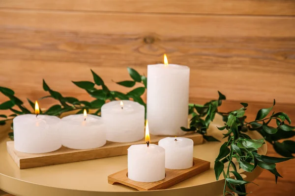 Burning candles with plant branches on table near wooden wall, closeup