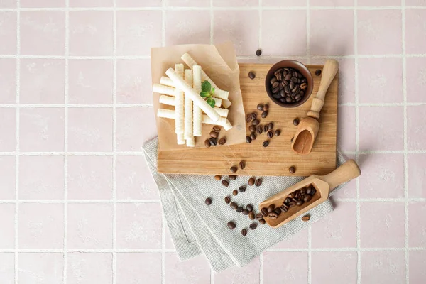 Board with delicious wafer rolls and coffee beans on pink tile background