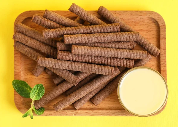 Board with delicious chocolate wafer rolls and condensed milk on yellow background