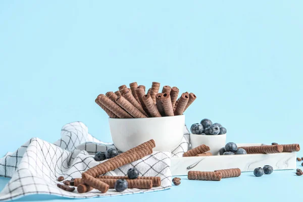 Bowl with delicious chocolate wafer rolls and blueberries on blue background