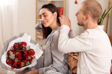 Young man putting necklace around his wife's neck in bedroom on Valentine's Day