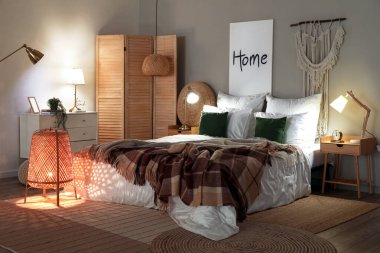 Interior of bedroom with brown checkered blankets on bed and glowing lamps late in evening