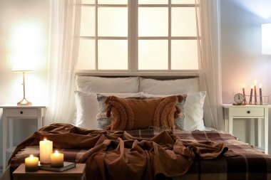 Interior of bedroom with brown checkered blankets on bed, burning candles and glowing lamps late in evening