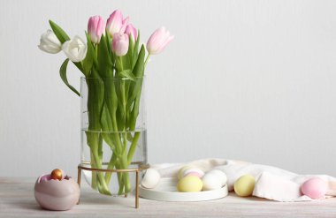 Vase with beautiful tulip flowers and Easter eggs on table near light wall