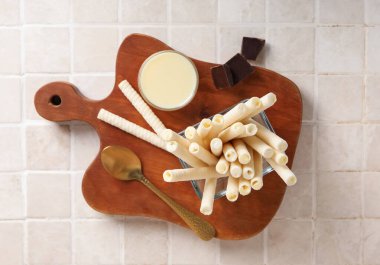 Board with delicious wafer rolls, condensed milk and chocolate pieces on tile background