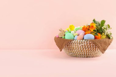 Wicker basket with Easter eggs, decor and flowers on color background