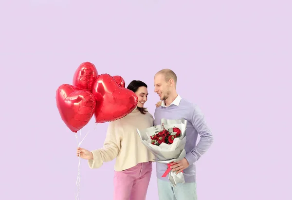 Young couple with bouquet of flowers and balloons on lilac background. Valentine's Day celebration