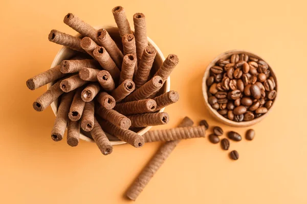 Bowls with delicious chocolate wafer rolls and coffee beans on orange background