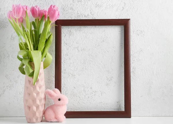 Vase with beautiful tulip flowers, empty picture frame and Easter bunny on light background