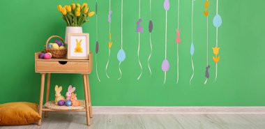 Beautiful tulips, basket with Easter eggs and bunnies on table near green wall in room