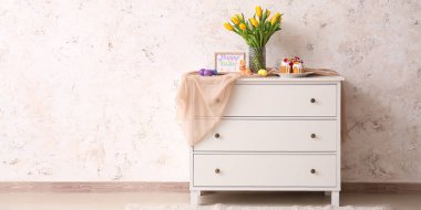 Vase with tulips, Easter cakes, bunny and eggs on chest of drawers near light wall in room
