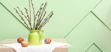 Pussy willow branches and Easter eggs on table near green wall