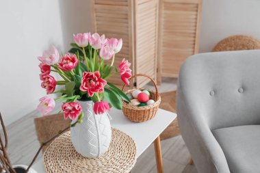 Vase with tulips and basket of Easter eggs on table in living room