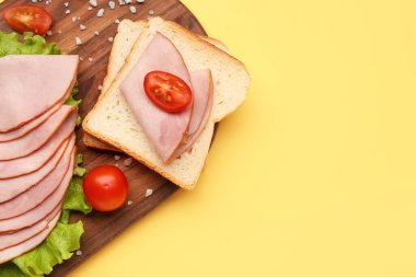 Board with slices of tasty ham, bread pieces, lettuce and tomatoes on yellow background