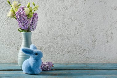 Vase with beautiful flowers and Easter bunny on blue wooden table near grey wall wall