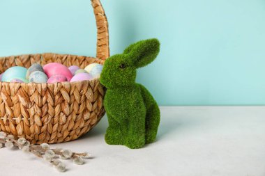Easter bunny, basket with painted eggs and pussy willow branches on table near blue wall