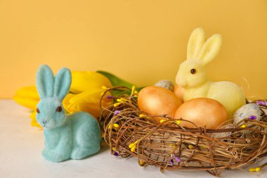 Easter bunnies, tulip flowers and nest with painted eggs on table against yellow background