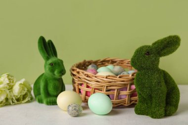 Easter bunnies and wicker basket with painted eggs on table against green background