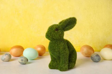 Easter bunny and painted eggs on table against yellow background