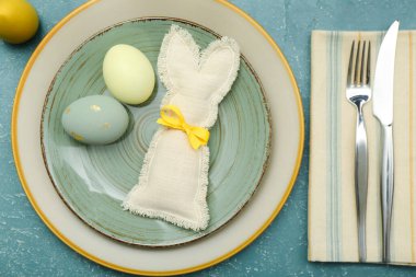 Table setting with Easter eggs, bunny and cutlery on blue background
