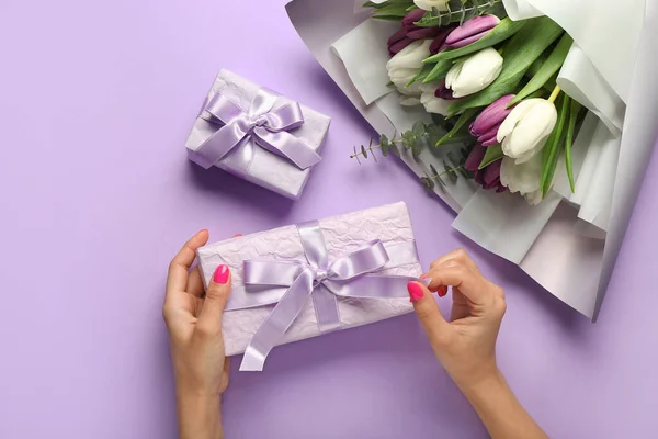 Female hands holding gift box and bouquet of tulip flowers on lilac background. Hello spring