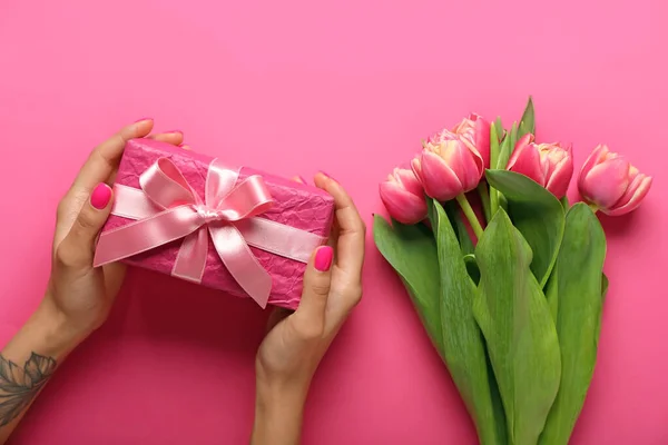 Female hands holding gift box and beautiful tulip flowers on pink background. Hello spring