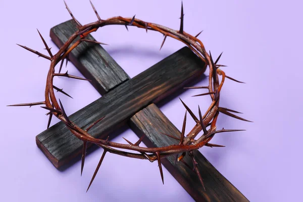 Wooden cross with crown of thorns on lilac background, closeup. Good Friday concept