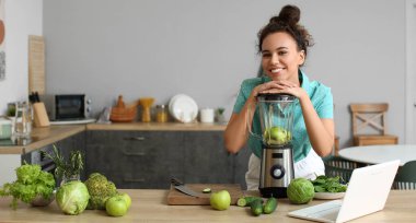 Young woman with modern blender making green smoothie in kitchen at home