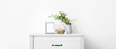 Blank photo frame, nest with Easter eggs, bunnies, green branches on chest of drawers near white wall. Banner for design