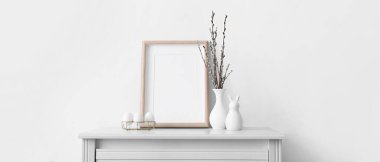 Blank photo frame, holder with Easter eggs, bunny and pussy willow branches on table near white wall. Banner for design