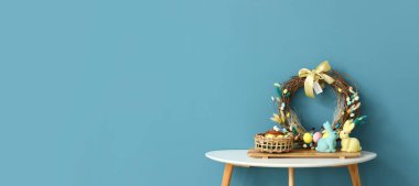 Basket with Easter eggs, cake, bunnies and beautiful Easter wreath on table near blue wall. Banner for design