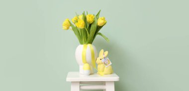 Beautiful yellow tulips, Easter egg and rabbit with greeting card on stool near light green wall