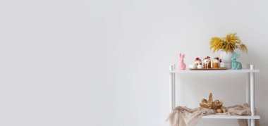 Rack with Easter cakes, eggs, bunnies and mimosa flowers near white wall. Banner for design