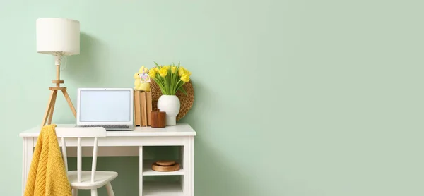Workplace with modern laptop, tulips and Easter rabbit with greeting card near light green wall. Banner for design