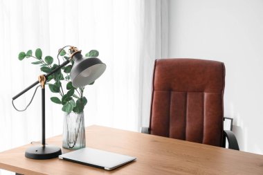 Vase with eucalyptus, laptop and lamp on table near light curtain in office