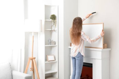 Young woman with blank frame nailing on light wall at home