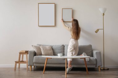 Young woman hanging blank frame on light wall at home, back view