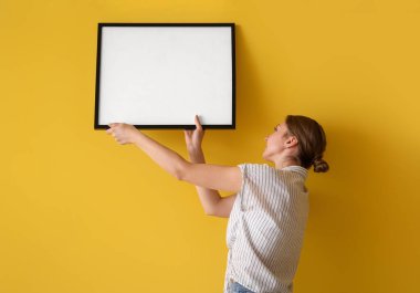 Young woman hanging blank frame on yellow wall at home