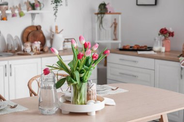 Vase with tulips, Easter eggs, cake and jug of water on dining table in kitchen